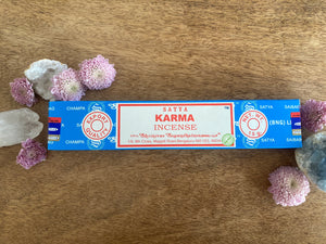 Satya Karma incense packaging on a wooden surface with flowers and crystals.