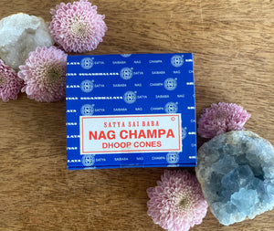 Box of Satya Nag Champa dhoop cones on a wooden surface with flowers and a crystal.